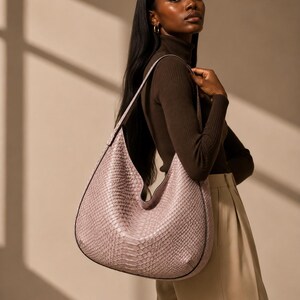 May include: A light pink, large, crescent-shaped handbag with a snakeskin texture. The bag has a black trim and a long shoulder strap. The model is wearing a brown turtleneck and beige trousers.