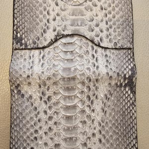 Puede incluir: Un tarjetero rectangular de piel de serpiente gris y blanca. El tarjetero tiene una abertura superior curvada y un borde cosido negro. El patrón de piel de serpiente es visible en toda la superficie.