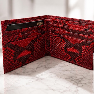 Puede incluir: Cartera roja abierta de dos pliegues con un patrón de piel de serpiente. La cartera tiene múltiples ranuras para tarjetas y un compartimento para billetes. Una tarjeta de crédito negra con "VISA" es visible dentro.