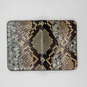 Puede incluir: Una cartera rectangular con un estampado de piel de serpiente. La cartera presenta una combinación de colores, incluyendo tonos de gris, negro y beige. El diseño de piel de serpiente se muestra prominentemente en la superficie.