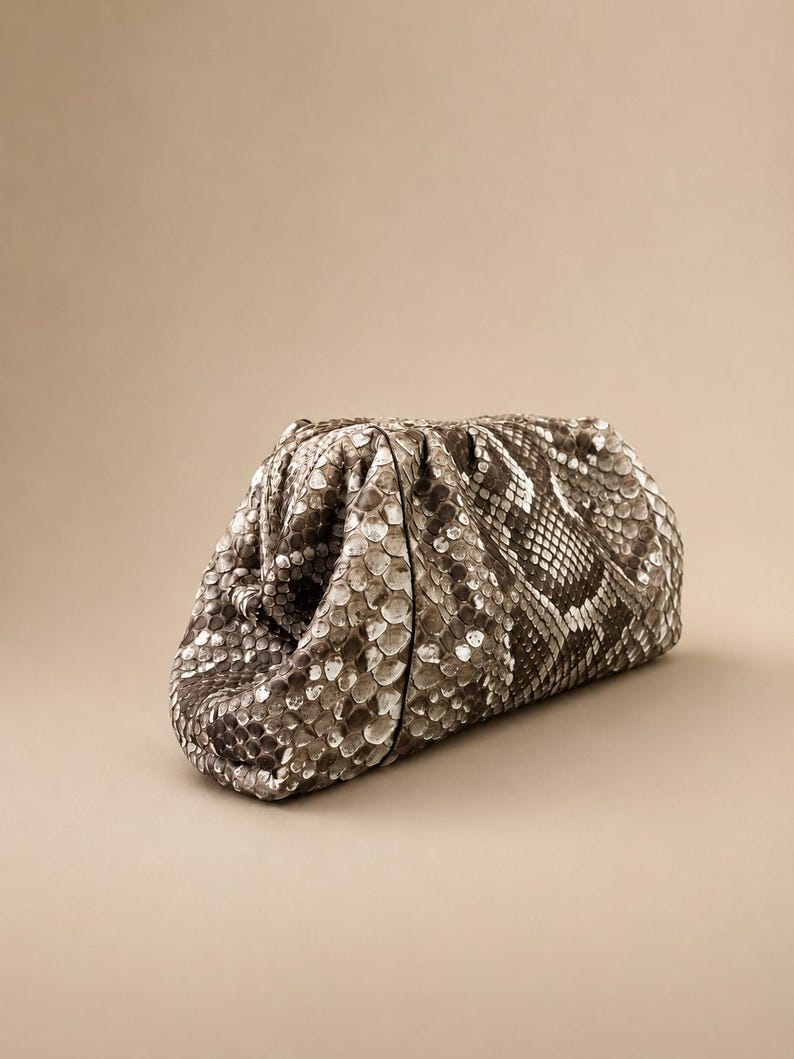 May include: A structured clutch bag featuring a textured snakeskin pattern in shades of gray, brown, and cream. The bag has a gathered, pouch-like silhouette with a soft, rounded shape and a concealed frame closure at the top.