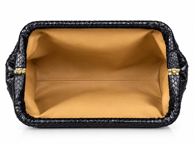 May include: An open rectangular clutch bag with a black exterior featuring a textured snakeskin pattern. The interior is lined with smooth tan leather. The frame of the bag is constructed with gold-toned metal hinges on both sides.