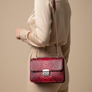 Puede incluir: Un bolso de hombro rectangular con un patrón de piel de serpiente en rojo y negro. El bolso tiene una correa de cadena de metal plateado y un cierre de presión rectangular plateado en la solapa frontal. La textura imita escamas de reptil.
