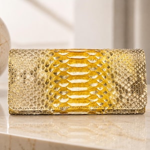 Könnte beinhalten: Eine rechteckige Clutch aus Schlangenleder mit einem Muster aus gelben und beigen Schuppen. Die Clutch ist geschlossen und scheint aus echtem Schlangenleder zu bestehen. Die Nähte sind an den Rändern sichtbar.