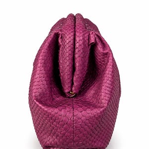 Pode incluir: Uma bolsa clutch magenta com um padr&atilde;o texturizado de pele de cobra. A bolsa apresenta um fecho de estrutura r&iacute;gida no topo, com um corpo arredondado e volumoso que se estreita em dire&ccedil;&atilde;o &agrave; base, exibindo uma textura de escamas.