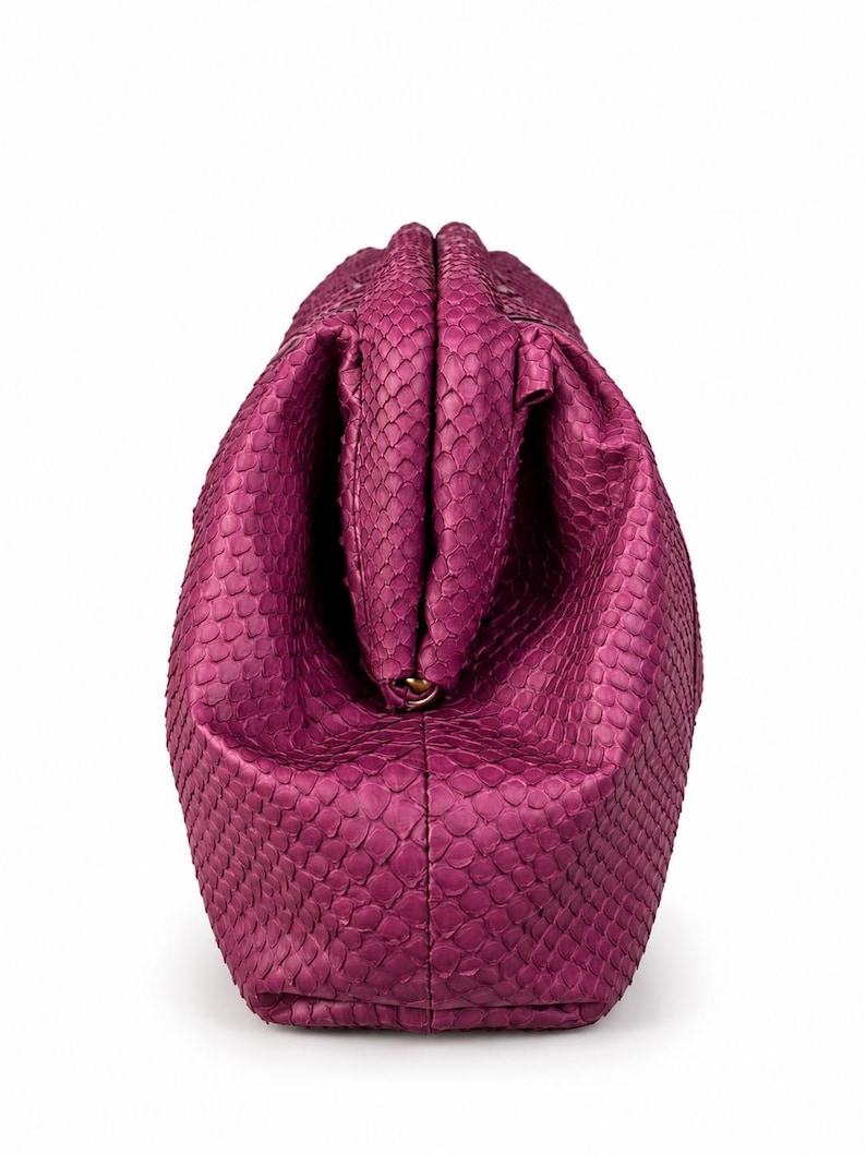 Pode incluir: Uma bolsa clutch magenta com um padr&atilde;o texturizado de pele de cobra. A bolsa apresenta um fecho de estrutura r&iacute;gida no topo, com um corpo arredondado e volumoso que se estreita em dire&ccedil;&atilde;o &agrave; base, exibindo uma textura de escamas.