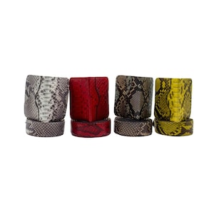 May include: Four square bracelets in a row, each with a different snakeskin pattern. The colours include grey, red, brown, and yellow. Each bracelet sits on a matching base, creating a cohesive design. The bracelets are likely made of leather.