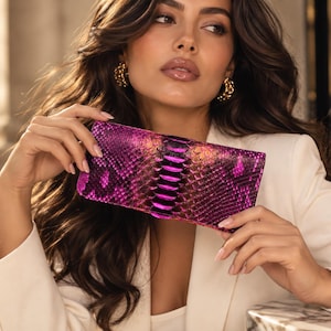 May include: A rectangular clutch bag featuring a vibrant magenta and black snakeskin-patterned texture. The accessory has a glossy, iridescent finish that shifts between deep purple and bright pink tones across its structured, elongated surface.