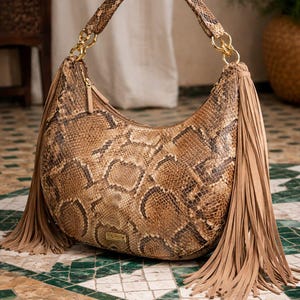 Könnte beinhalten: Eine braun-beige Handtasche mit Schlangenmuster, geschwungener Form und langem Henkel. Die Tasche hat goldfarbene Kettenakzente und lange, beige Fransen auf jeder Seite. Die Tasche steht auf einem grün-weiß gefliesten Boden.