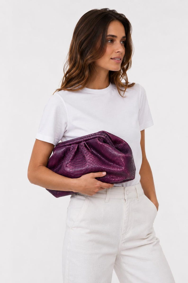 May include: A deep purple clutch bag with a textured snakeskin pattern. The bag features a gathered, soft-structured design with a hinged frame closure, providing a voluminous silhouette that is held horizontally against the midsection.
