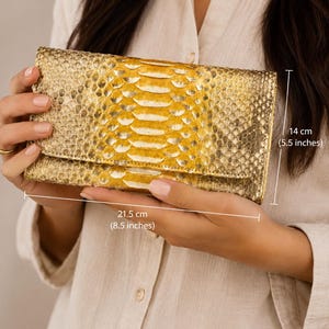 Könnte beinhalten: Eine rechteckige goldene Clutch mit strukturiertem Schlangenledermuster und einem Mittelstreifen aus gelbem Fell. Die Clutch ist 21,5 cm breit und 14 cm hoch, verfügt über einen Klappverschluss und eine metallische Oberfläche auf der Außenseite.
