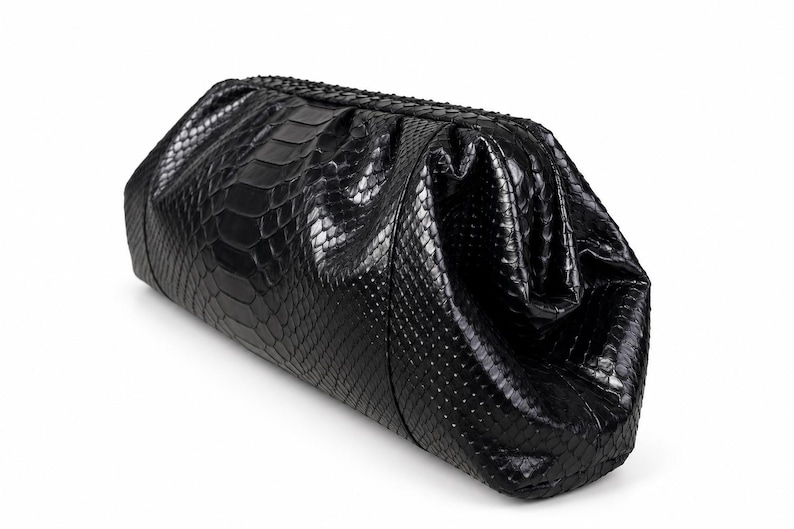 May include: A black clutch bag with a textured snakeskin pattern. The bag features a structured, elongated rectangular shape with a gathered top closure and a glossy finish that highlights the intricate scale details across the entire surface.