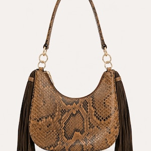 Peut inclure: Un sac à bandoulière en forme de croissant avec un motif peau de serpent marron et noir. Le sac comprend une anse unique fixée par des anneaux métalliques dorés et de longues franges en suédine marron foncé sur les côtés de la fermeture.