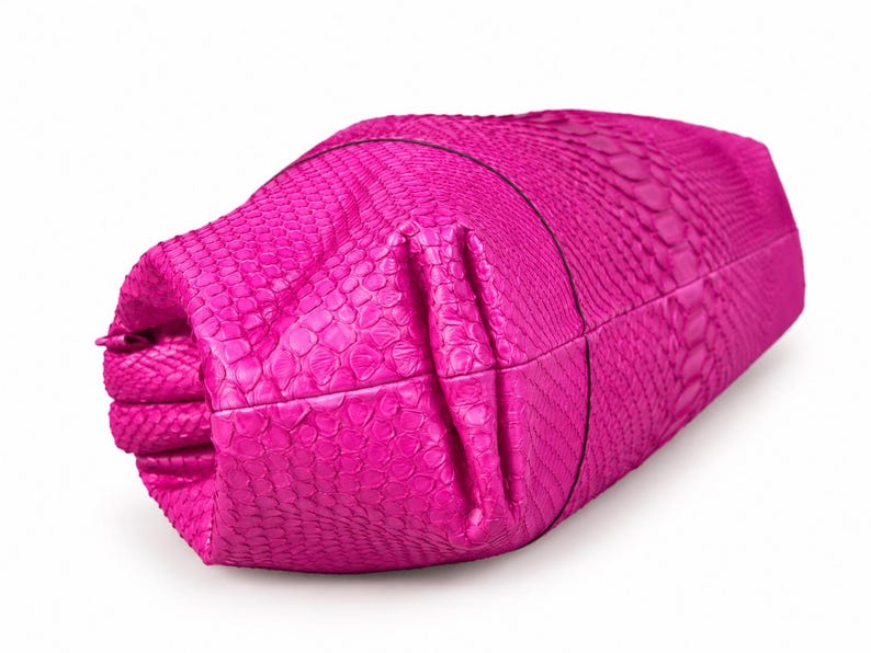 May include: A bright fuchsia clutch bag featuring a textured snakeskin pattern. The bag has a folded, structured design with visible seams and a smooth, consistent color throughout the exterior surface, showcasing a detailed scale-like texture.