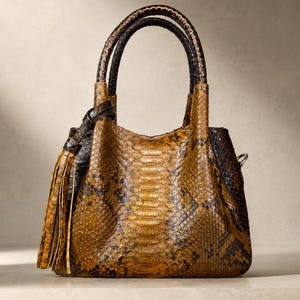 May include: A structured handbag featuring a textured snakeskin pattern in shades of golden brown and dark charcoal. The bag includes two rounded top handles, a side tassel detail, and a curved top opening, showcasing a detailed scale texture.