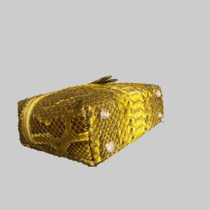May include: A rectangular pouch featuring a metallic gold and yellow snakeskin-textured exterior. The bag includes a matching gold zipper closure along the top edge and four small, clear protective feet attached to the bottom corners.