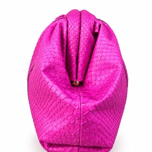 May include: A vibrant hot pink clutch bag featuring a textured snakeskin pattern. The bag has a structured, rounded shape with a central kiss-lock closure mechanism made of gold-toned metal, visible at the top center of the folded opening.