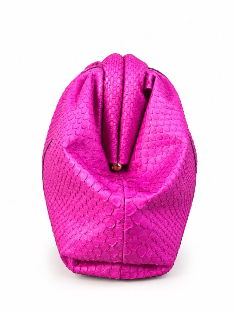 May include: A vibrant hot pink clutch bag featuring a textured snakeskin pattern. The bag has a structured, rounded shape with a central kiss-lock closure mechanism made of gold-toned metal, visible at the top center of the folded opening.