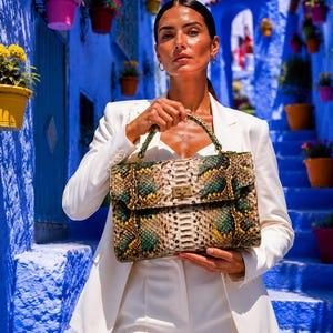 May include: A rectangular handbag with a snakeskin pattern in shades of green, brown, and yellow. The bag has a gold-coloured clasp and a green handle. The person is wearing a white blazer and trousers. The background is a blue alleyway with flower pots.