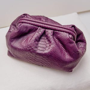 May include: A deep purple clutch bag featuring a textured snakeskin pattern. The bag has a soft, gathered, pouch-like silhouette with a structured frame closure at the top, creating a pleated effect across the front and sides of the exterior.