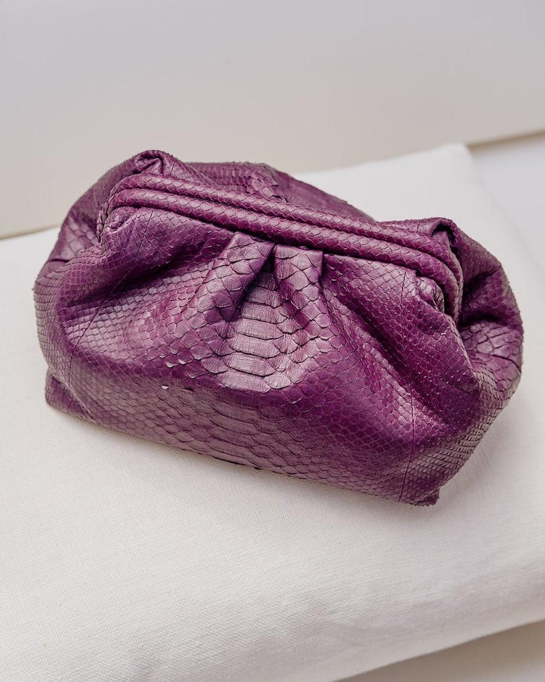 May include: A deep purple clutch bag featuring a textured snakeskin pattern. The bag has a soft, gathered, pouch-like silhouette with a structured frame closure at the top, creating a pleated effect across the front and sides of the exterior.