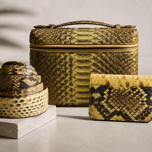May include: A set of accessories featuring a rectangular vanity case, a coiled belt, and a small wallet, all crafted with a yellow and black snakeskin pattern. The vanity case has a top handle and a zipper closure, while the wallet is rectangular.