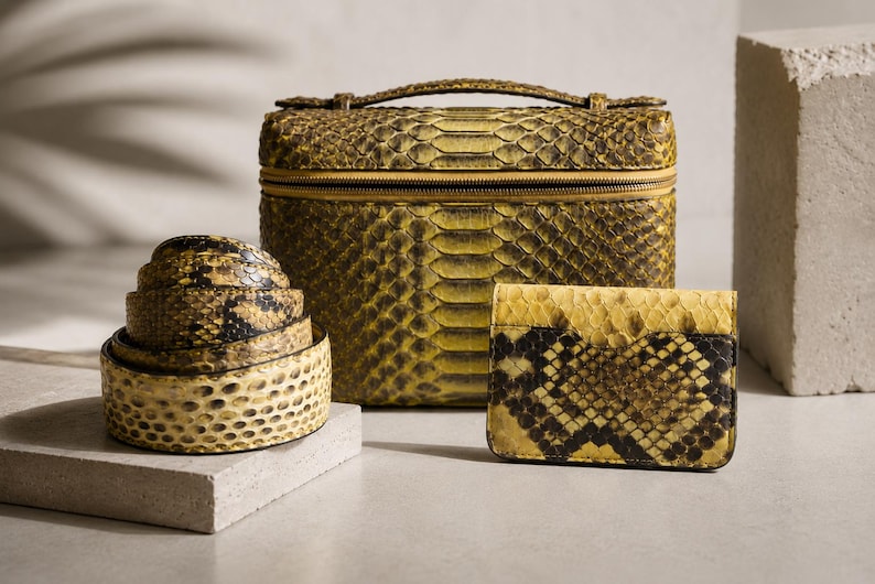 May include: A set of accessories featuring a rectangular vanity case, a coiled belt, and a small wallet, all crafted with a yellow and black snakeskin pattern. The vanity case has a top handle and a zipper closure, while the wallet is rectangular.