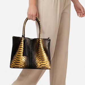 May include: A black and gold tote bag with a snakeskin pattern. The bag has a structured shape, a tassel, and braided handles. The bag is held by a person wearing beige trousers and shoes.