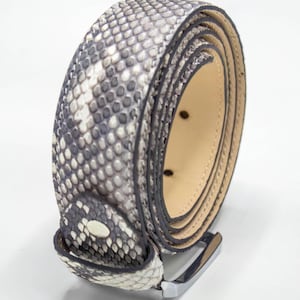 Può includere: Una cintura arrotolata con un motivo a pelle di serpente nei toni del grigio e del bianco. La cintura ha una fibbia color argento e un interno color beige chiaro. Il motivo a pelle di serpente è dettagliato e strutturato.