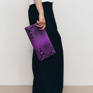 Könnte beinhalten: Eine rechteckige Clutch mit einem leuchtend violetten und schwarzen Schlangenledermuster. Das Accessoire hat ein schmales, flaches Profil mit einem spitz zulaufenden Klappenverschluss und zeigt eine detaillierte Reptilienstruktur.
