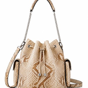 Könnte beinhalten: Beige Eimer-Tasche mit Schlangenmuster. Die Tasche hat einen Kordelzugverschluss, zwei Seitentaschen, einen oberen Griff und einen silbernen Kettenriemen. Die Tasche ist ca. 25 cm hoch und 20 cm breit.