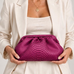Pode incluir: Uma bolsa clutch magenta vibrante com um padr&atilde;o texturizado de pele de cobra. A bolsa tem uma silhueta macia, arredondada e tipo bolsa com um fecho franzido no topo, exibindo uma textura de superf&iacute;cie escamosa em tom rosa profundo.
