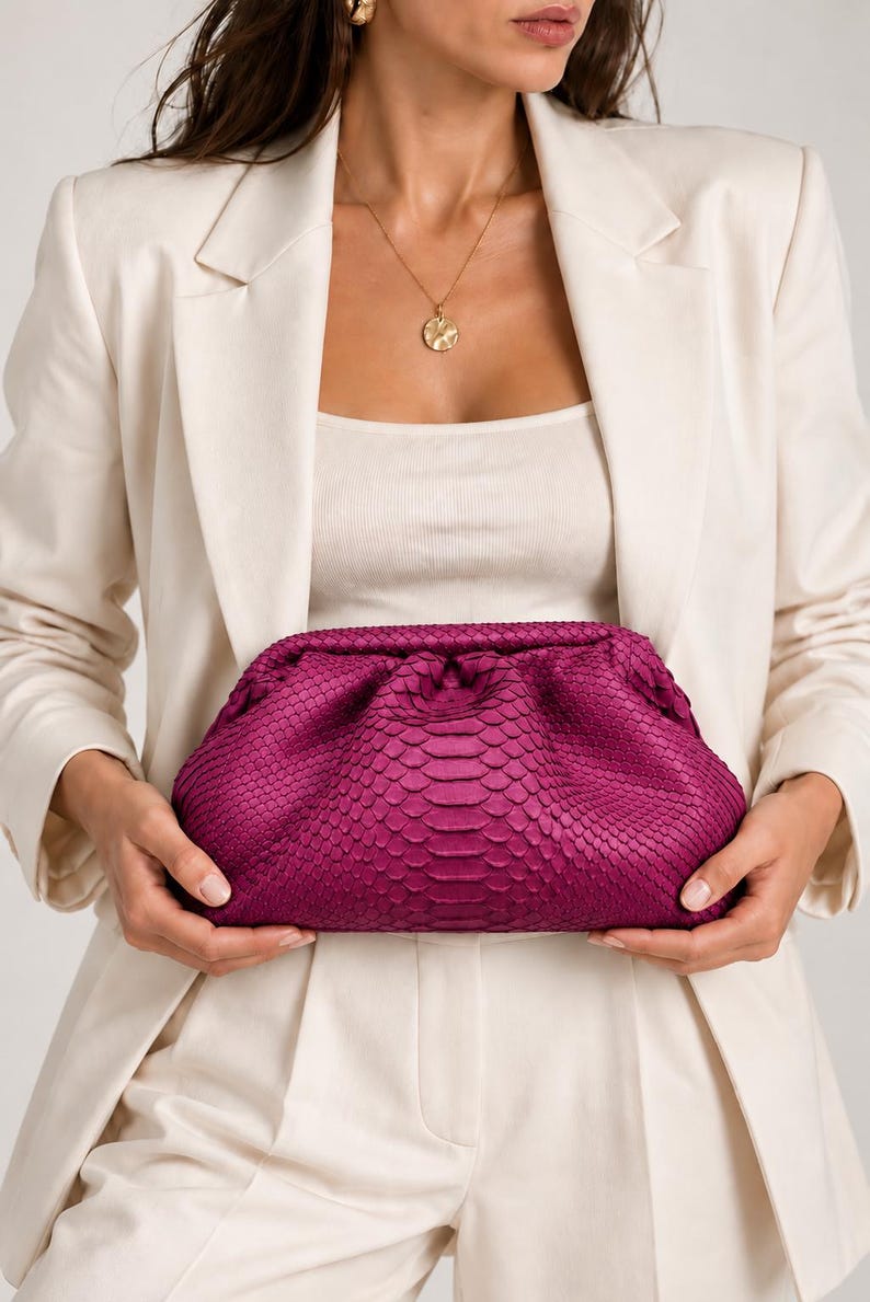 Pode incluir: Uma bolsa clutch magenta vibrante com um padr&atilde;o texturizado de pele de cobra. A bolsa tem uma silhueta macia, arredondada e tipo bolsa com um fecho franzido no topo, exibindo uma textura de superf&iacute;cie escamosa em tom rosa profundo.