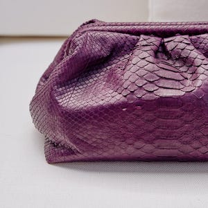 May include: A deep purple clutch bag featuring a textured snakeskin pattern. The bag has a soft, gathered, pouch-like silhouette with a structured frame opening at the top, showcasing a detailed scale texture across the entire exterior surface.
