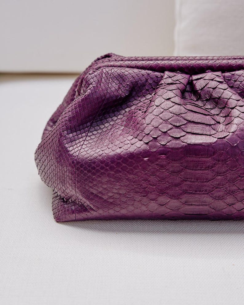 May include: A deep purple clutch bag featuring a textured snakeskin pattern. The bag has a soft, gathered, pouch-like silhouette with a structured frame opening at the top, showcasing a detailed scale texture across the entire exterior surface.