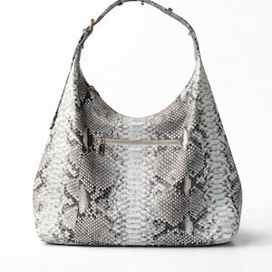 Può includere: Una grande borsa a spalla bianca e grigia con motivo a pelle di serpente e una sola tracolla. La borsa ha una forma curva e una chiusura con cerniera. L'hardware è color oro.