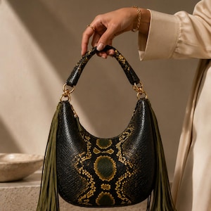 Puede incluir: Bolso de mano negro y dorado con estampado de piel de serpiente y borlas verde oliva. El bolso en forma de media luna tiene un asa curva y herrajes dorados. El bolso lo sostiene una persona.