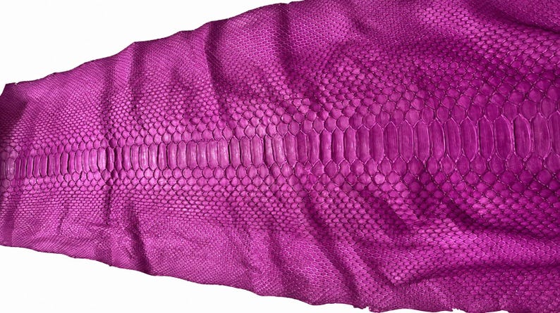 May include: A vibrant magenta snakeskin leather hide featuring a distinct central row of large, elongated scales flanked by smaller, uniform hexagonal scales on both sides, showcasing a natural, textured pattern throughout the entire surface.