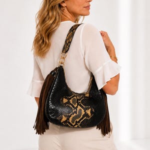 Könnte beinhalten: Eine Hobo-Schultertasche mit schwarz-beigem Schlangenledermuster, einem breiten passenden Schulterriemen und langen, dunkelbraunen Wildlederfransen an beiden Seiten. Die Tasche verfügt über ein goldfarbenes Kettenglied und einen Reißverschluss.