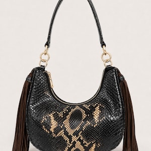 May include: A crescent-shaped shoulder bag featuring a black and tan snakeskin-patterned exterior. The bag includes a single top handle attached with gold-toned metal rings, a top zip closure, and long dark brown suede fringe tassels on both sides.