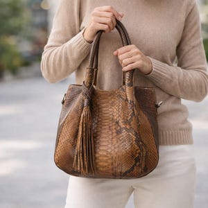 Könnte beinhalten: Eine braune Handtasche mit Schlangenmuster und Quaste. Die Tasche hat eine strukturierte Form mit einem oberen Griff und einem Schultergurt. Die Tasche wird von einer Person gehalten, die einen beigen Pullover und eine weiße Hose trägt.