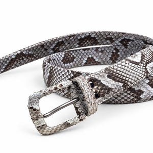 Pode incluir: Um cinto com um padrão texturizado de pele de cobra em tons de cinza, branco e castanho escuro. O cinto inclui uma fivela retangular a condizer revestida a pele de cobra com um pino de metal fino e escuro e uma presilha.
