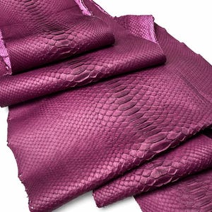 Pode incluir: Uma pe&ccedil;a dobrada de couro de cobra na cor magenta, apresentando um padr&atilde;o de escamas natural e distinto. O material exibe um tom roxo profundo e vibrante com uma superf&iacute;cie texturizada que destaca a disposi&ccedil;&atilde;o org&acirc;nica das escamas individuais.