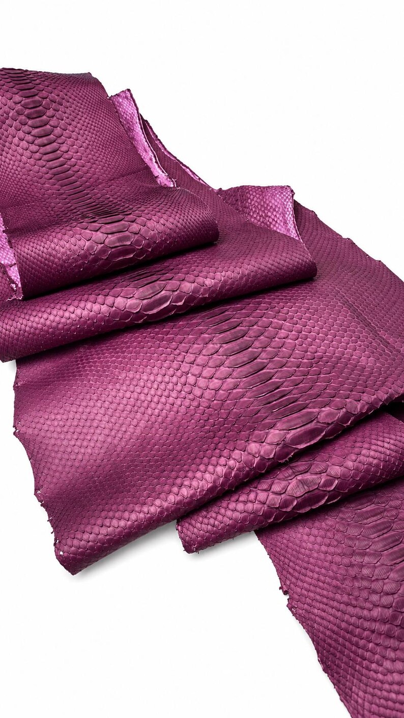 Pode incluir: Uma pe&ccedil;a dobrada de couro de cobra na cor magenta, apresentando um padr&atilde;o de escamas natural e distinto. O material exibe um tom roxo profundo e vibrante com uma superf&iacute;cie texturizada que destaca a disposi&ccedil;&atilde;o org&acirc;nica das escamas individuais.