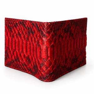 May include: A red bi-fold wallet made of textured material, possibly leather or a similar material. The wallet has a snakeskin pattern and a dark red color with black accents. The wallet is shown open, displaying its interior.