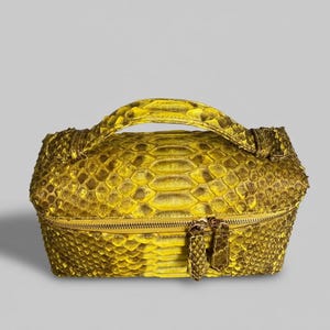 May include: A rectangular vanity case featuring a vibrant yellow and brown snakeskin pattern. The bag includes a top handle, a structured boxy silhouette, and a horizontal zipper closure with two matching snakeskin-textured zipper pulls.
