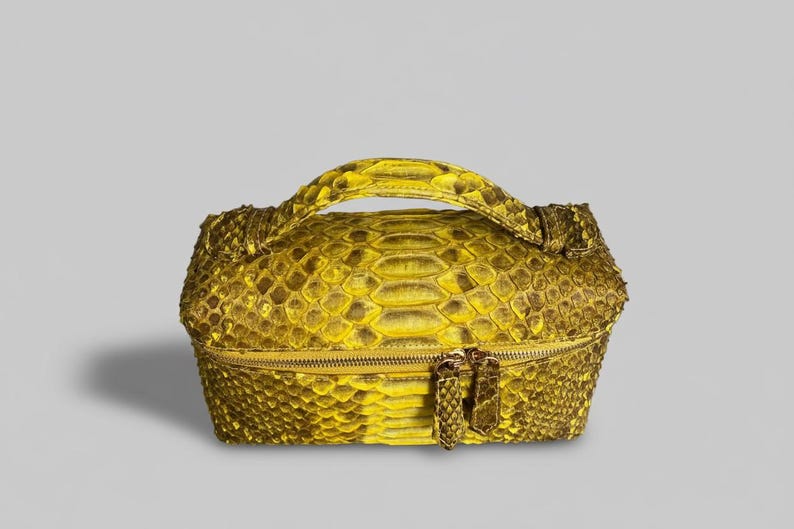 May include: A rectangular vanity case featuring a vibrant yellow and brown snakeskin pattern. The bag includes a top handle, a structured boxy silhouette, and a horizontal zipper closure with two matching snakeskin-textured zipper pulls.