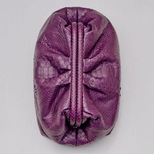 May include: A deep purple clutch bag with a textured snakeskin pattern. The bag features a structured, vertical central frame closure that divides the body into two gathered, rounded sections, creating a soft, voluminous, and pleated appearance.