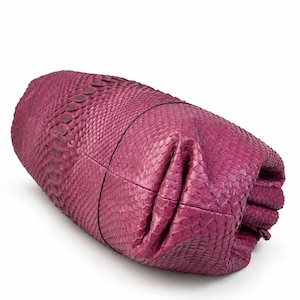 Pode incluir: Uma bolsa clutch na cor magenta com um padr&atilde;o texturizado semelhante a escamas em toda a sua superf&iacute;cie. A bolsa tem um formato cil&iacute;ndrico e dobrado com costuras vis&iacute;veis e uma estrutura macia e flex&iacute;vel que parece ser de pele ex&oacute;tica.