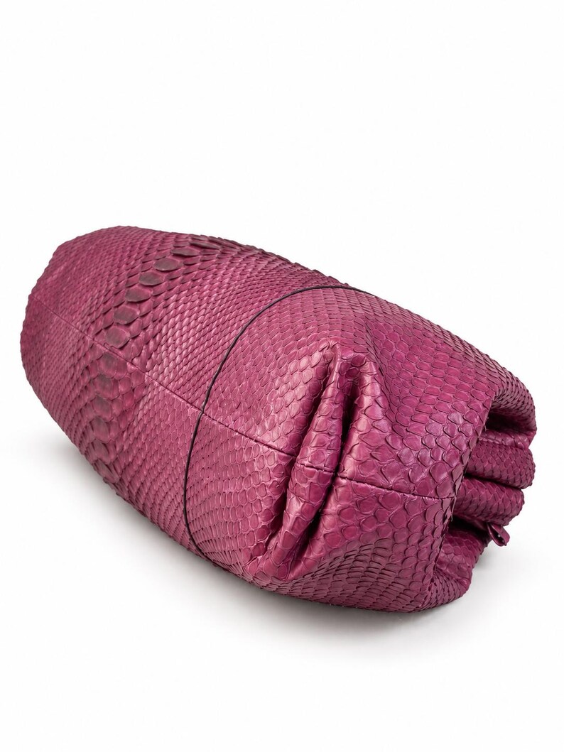 Pode incluir: Uma bolsa clutch na cor magenta com um padr&atilde;o texturizado semelhante a escamas em toda a sua superf&iacute;cie. A bolsa tem um formato cil&iacute;ndrico e dobrado com costuras vis&iacute;veis e uma estrutura macia e flex&iacute;vel que parece ser de pele ex&oacute;tica.